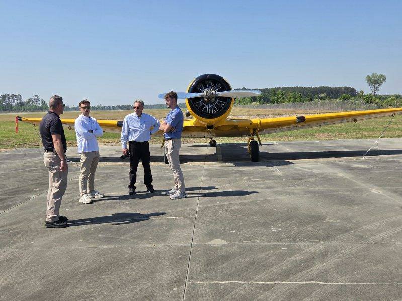 Four men stand talking on an airstrip in front of a yellow propeller airplane, with clear skies and a distant treeline in the background.