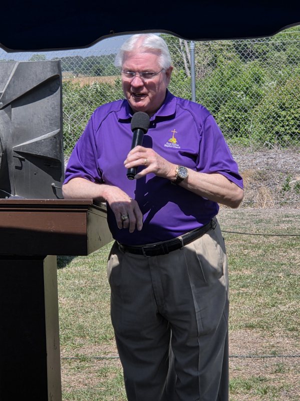 older man in a purple shirt speaks into a microphone outdoors, standing beside a podium with a fence and greenery in the background.