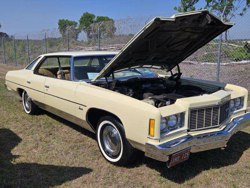 A vintage cream-colored car with its hood open is parked on grass near a fence, displaying the engine at an outdoor event.