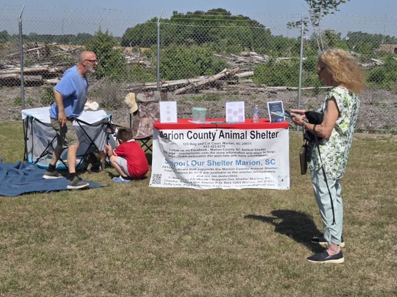 People stand near a Marion County Animal Shelter booth outdoors with pamphlets and a banner displayed, while dogs and strollers are visible on a sunny day.