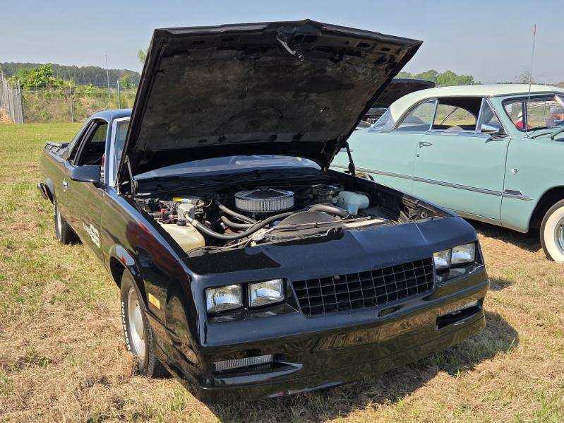 Black classic car with hood open, showing engine. Parked outdoors on grass next to a light blue vintage car. Clear sunny day.