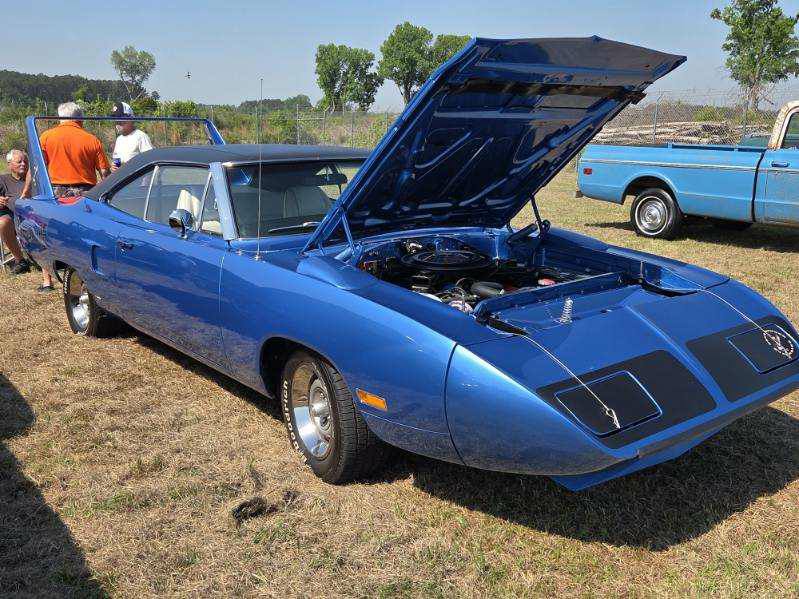 Blue classic muscle car with its hood open parked on grass at a car show, another blue car and people are visible in the background.