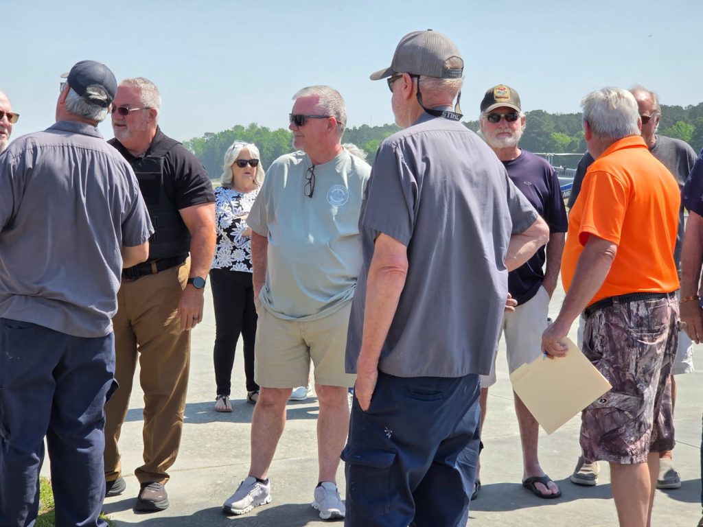 A group of people standing outdoors on a sunny day, some wearing sunglasses and hats, engaged in conversation.