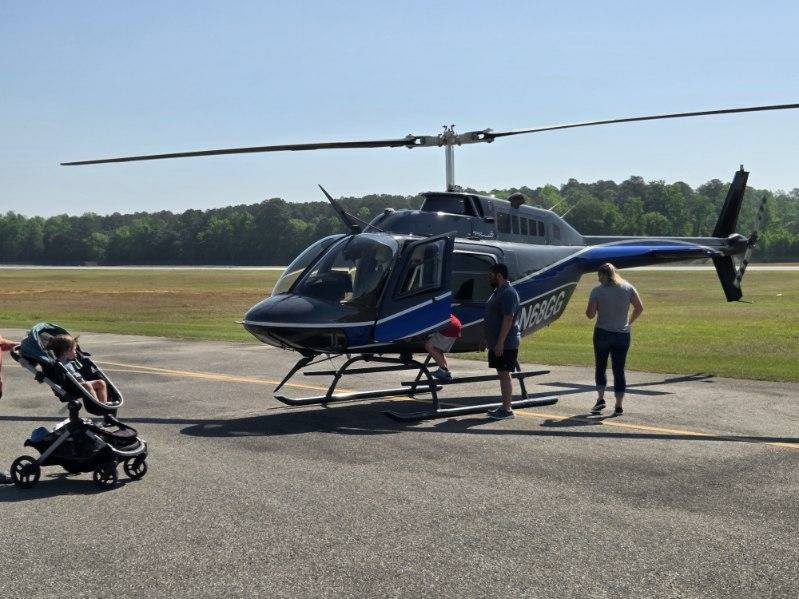 A blue and black helicopter on a tarmac with three people nearby, one in a wheelchair. Trees and open field in the background.