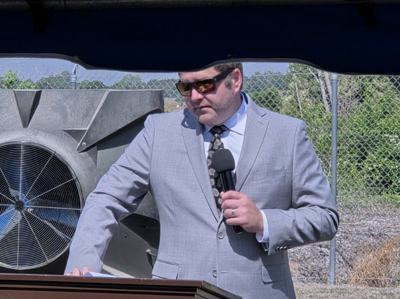 Man in a gray suit and sunglasses holding a microphone, standing outdoors at a podium with industrial equipment and greenery in the background.