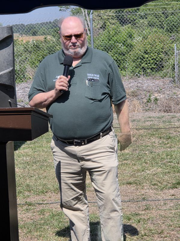 Man standing outdoors holding a microphone, wearing sunglasses, a green polo shirt, and khaki pants, speaking at a podium with a fence and greenery in the background.