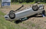 A silver sedan is overturned on its roof in a grassy ditch near a road, with trees and a sign visible in the background.