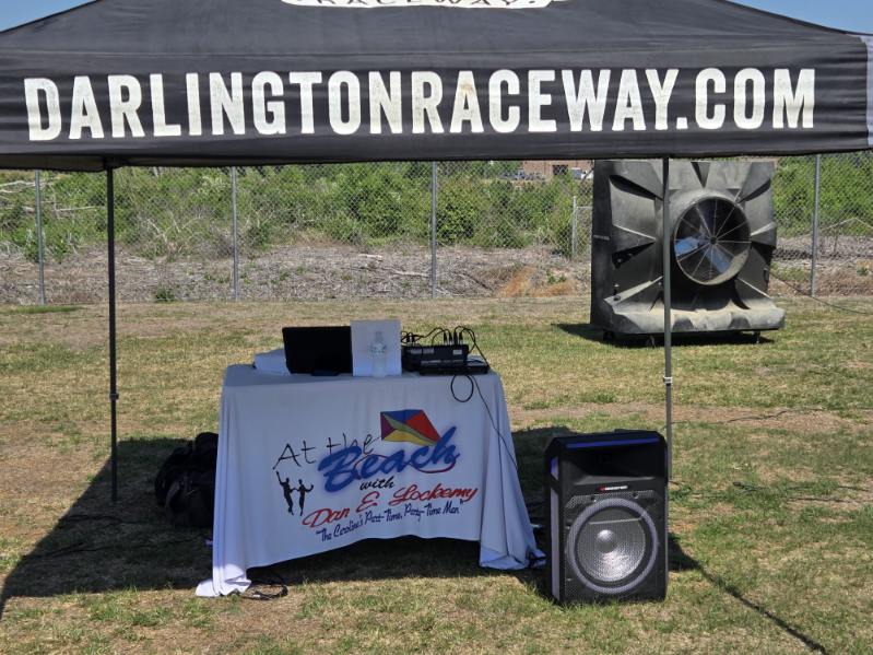 A booth setup outdoors with audio equipment on a table covered by a cloth, beneath a canopy labeled &ldquo;ARLINGTONRACEWAY.COM.&rdquo;