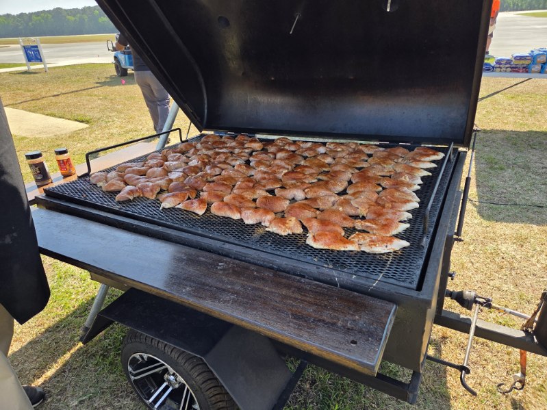 Large outdoor grill filled with rows of seasoned chicken pieces cooking, with the grill lid open on a sunny day.