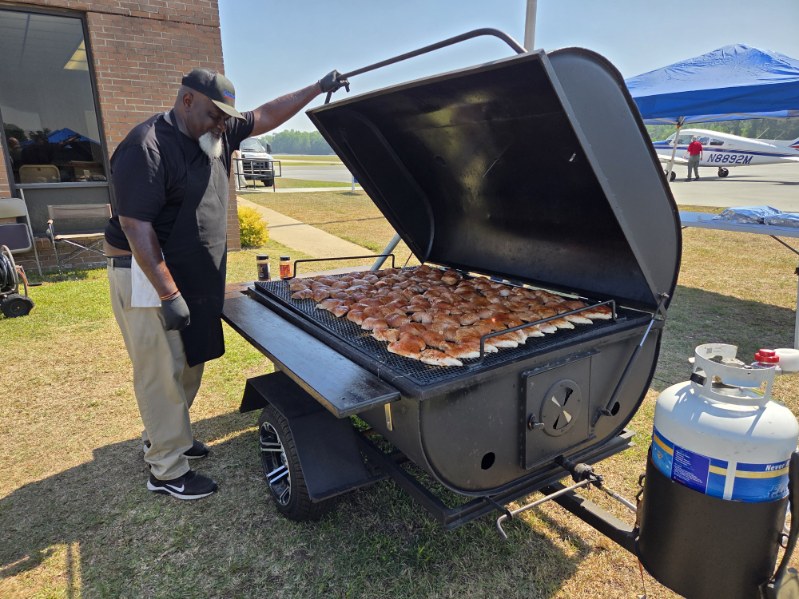 Man grilling a large quantity of chicken on an outdoor barbecue smoker grill, wearing gloves and apron, under sunny skies.