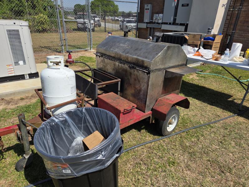 Outdoor metal barbecue grill on wheels with attached propane tank, near a trash bin lined with a plastic bag, set on grass beside a fenced area and buildings.