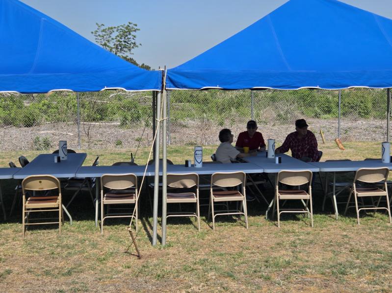 Three people sit under a blue tent at a long table with folding chairs outdoors, with grassy field and trees in the background.