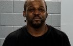 Man with a beard wearing a black shirt stands in front of a gray cinder block wall, looking at the camera.