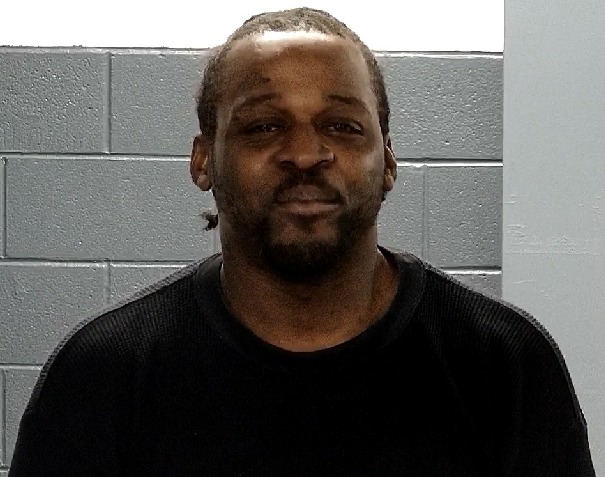 Man with a beard wearing a black shirt stands in front of a gray cinder block wall, looking at the camera.