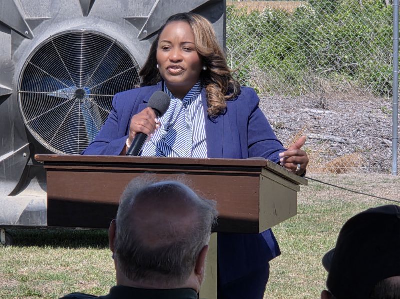 A woman in a blue suit speaks at a podium outdoors, holding a microphone, with people seated in front of her and a large fan structure behind.