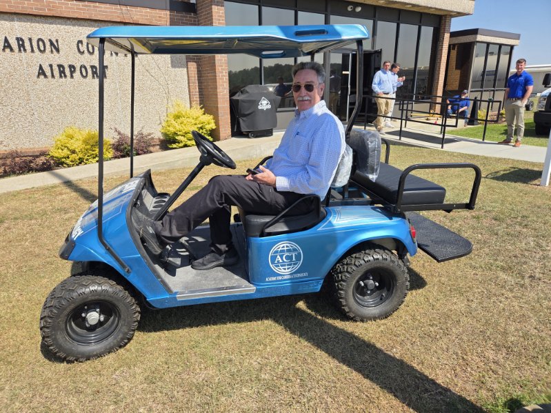 A man wearing sunglasses sits in a blue golf cart outside a building on a sunny day, with another person standing in the background.