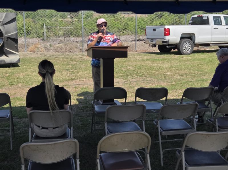 A person speaks at a podium outdoors to a mostly empty audience, with a few people seated and a white truck parked nearby.