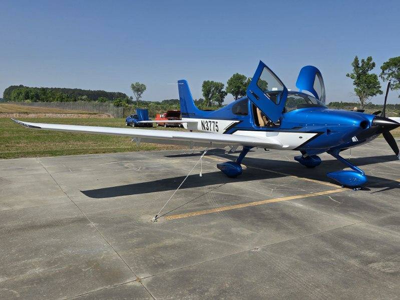 A small blue and white single-engine airplane parked on an airfield, with its canopy open and trees visible in the background under a clear sky.
