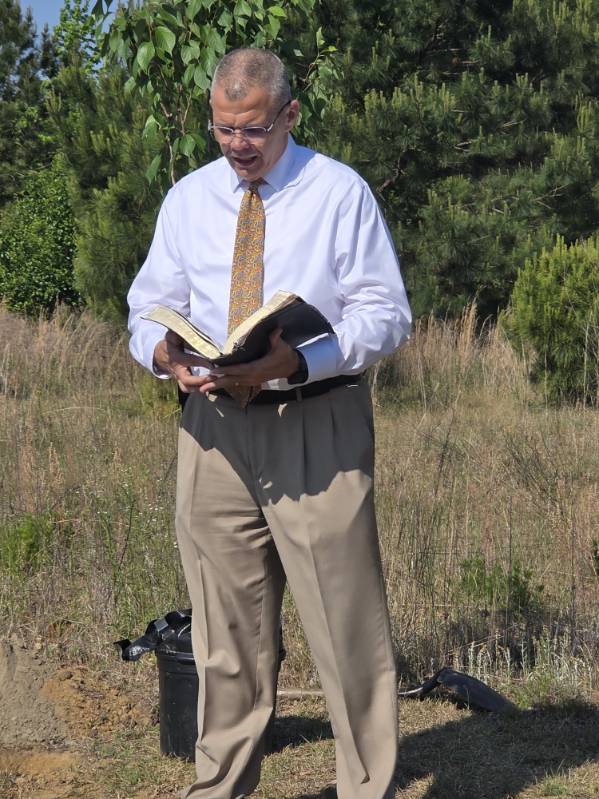 Man in dress shirt and tie standing outdoors, reading from a large open book with trees and tall grass in the background.