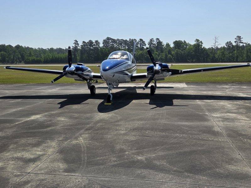 A twin-engine airplane is parked on a runway, with trees and clear skies in the background.