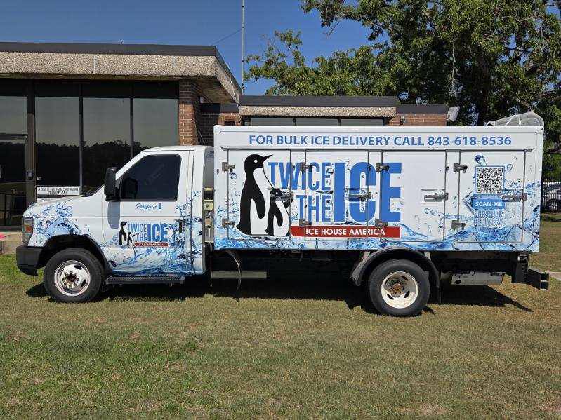 A delivery truck with "Twice the Ice" branding, penguin graphics, and contact info for bulk ice delivery, parked on grass beside a building.