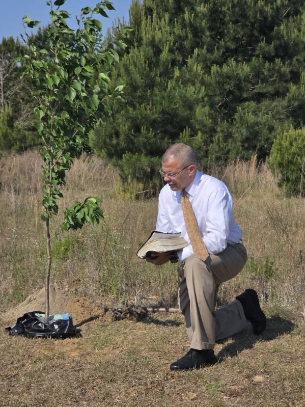 A man in a suit kneels outdoors beside a young tree, holding an open book, with grass and trees in the background.
