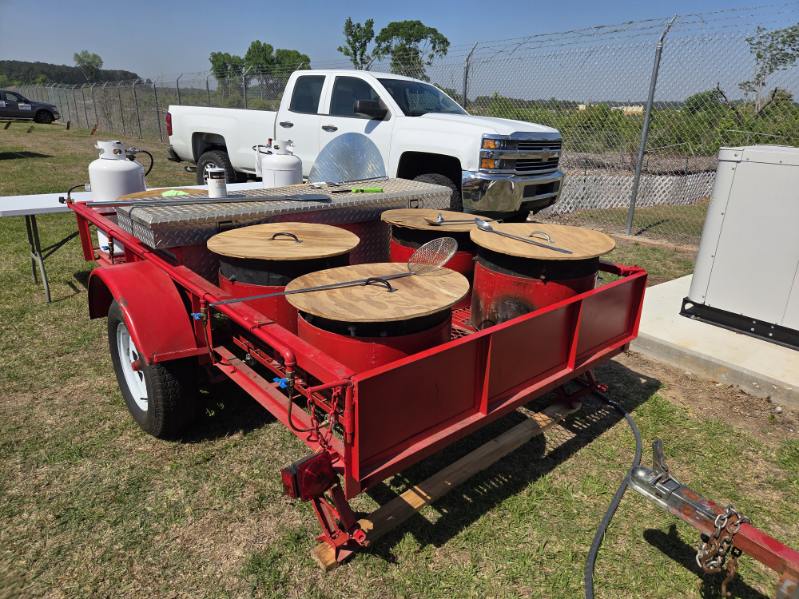 A red trailer with four round grills or smokers, parked outdoors near a white pickup truck on a grassy area.