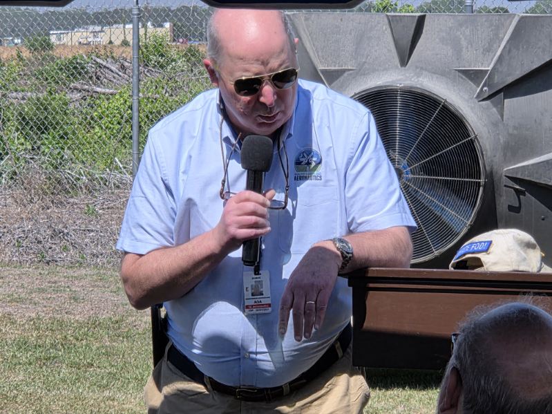 A man wearing sunglasses and a light blue shirt speaks into a microphone outdoors, standing near industrial equipment and a chain-link fence.