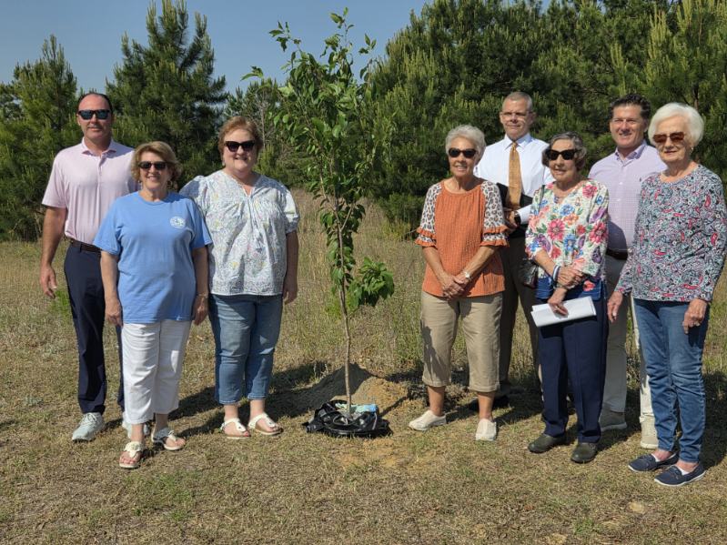 Seven adults stand outdoors around a newly planted tree on a sunny day, with green trees and blue sky in the background.