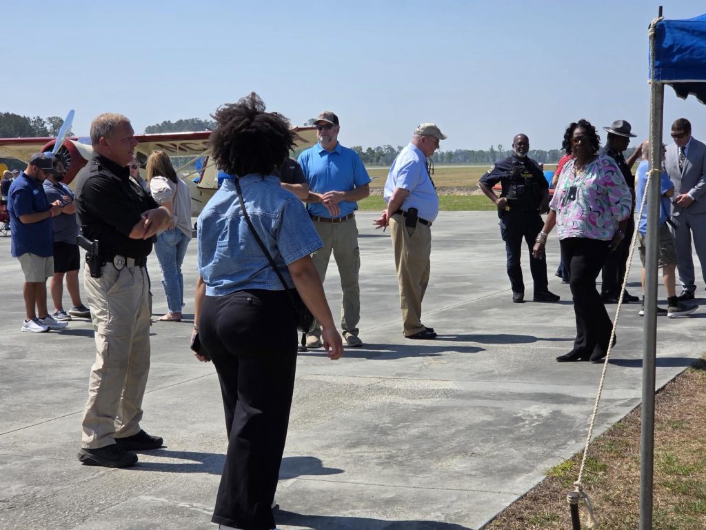A group of people stands outside on a paved area, engaged in conversation, with a clear sky in the background.