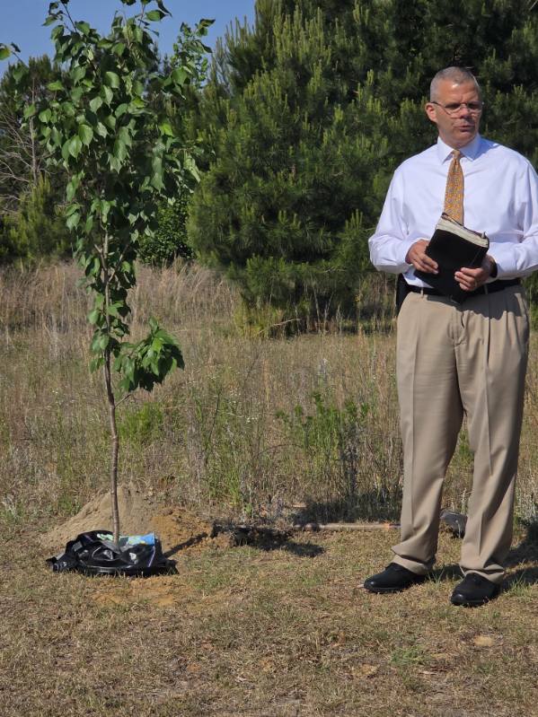 A man in dress clothes stands outdoors next to a newly planted tree, holding a book or folder, with grassy field and trees in the background.