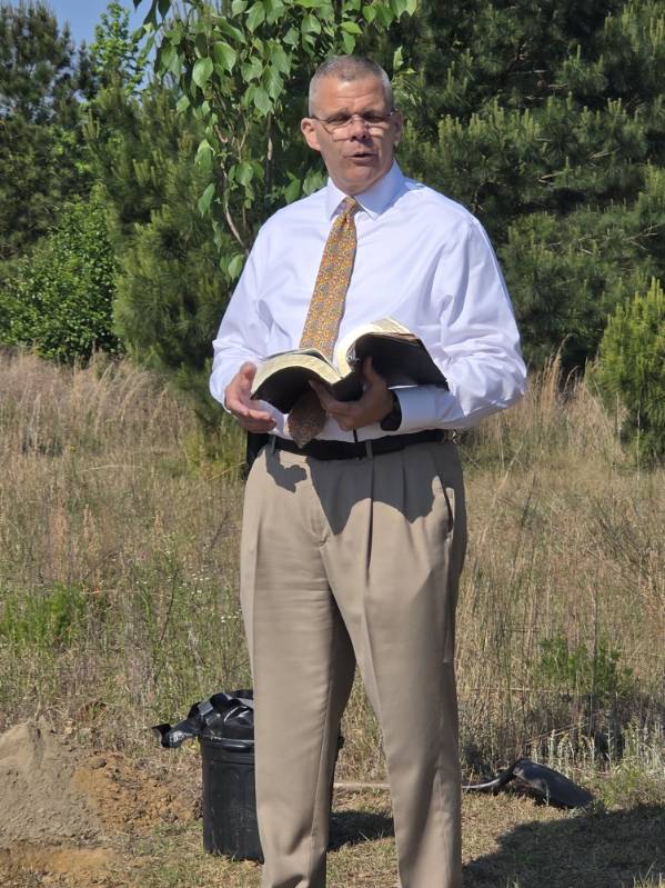 A man in a white shirt and tie stands outdoors, holding an open book, possibly giving a speech or sermon, surrounded by grass and trees.