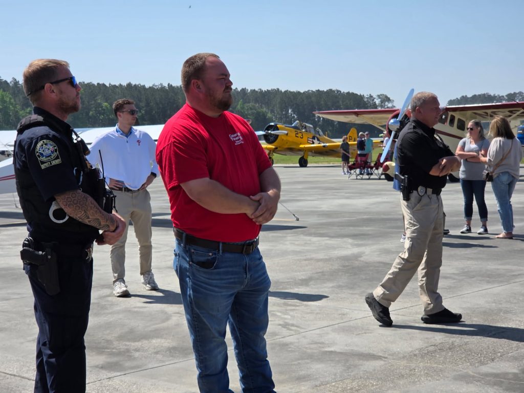 Several men stand on an airport tarmac, some wearing uniforms, with airplanes and clear skies in the background.