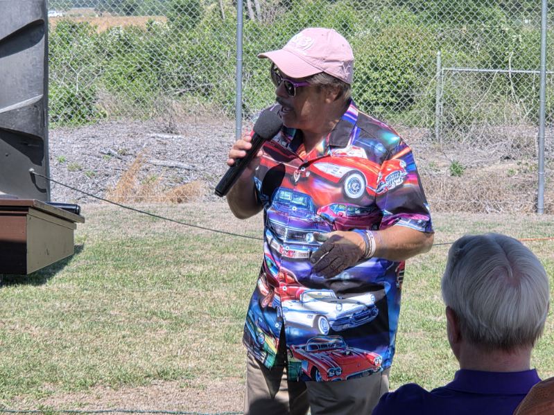 Person wearing a colorful shirt and pink cap speaks into a microphone outdoors, holding a glass, with greenery and a fence in the background.