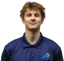 Young man with light brown hair wearing a navy blue collared shirt with a cartoon logo, standing and smiling slightly against a plain white background.