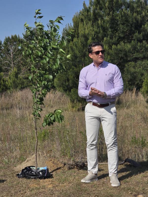 A man in a purple shirt and sunglasses stands outdoors next to a newly planted tree, with grass and pine trees in the background.