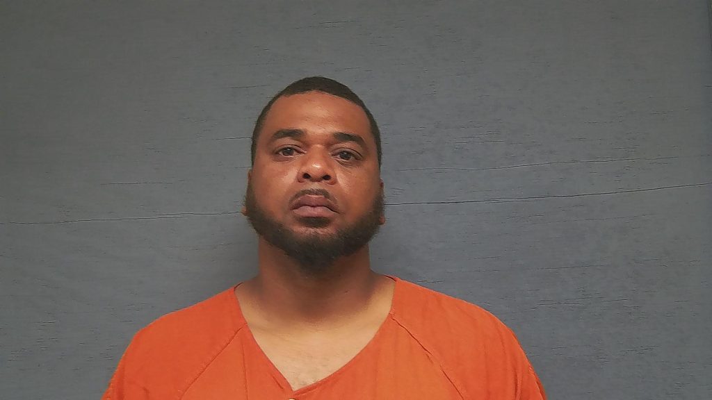 A man with short hair and a beard wearing an orange prison jumpsuit stands in front of a plain gray background.