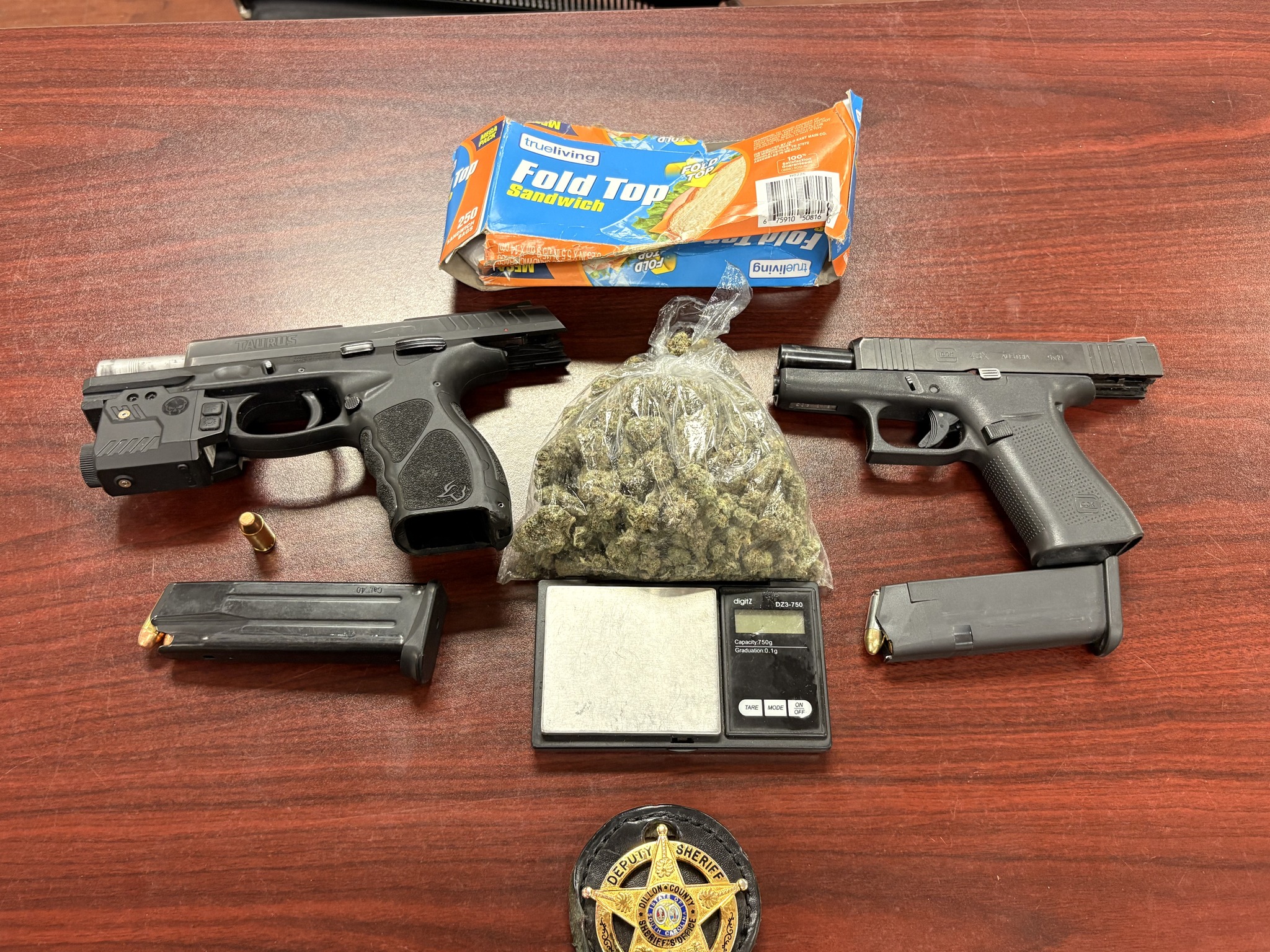 Two handguns, magazines, a bag of marijuana, a digital scale, sandwich bags, a sheriff’s badge, and bullets laid out on a wooden table.