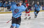 Ice hockey player in a blue uniform celebrates on the ice during a game, teammates and opponents in the background, with fans watching from the stands.