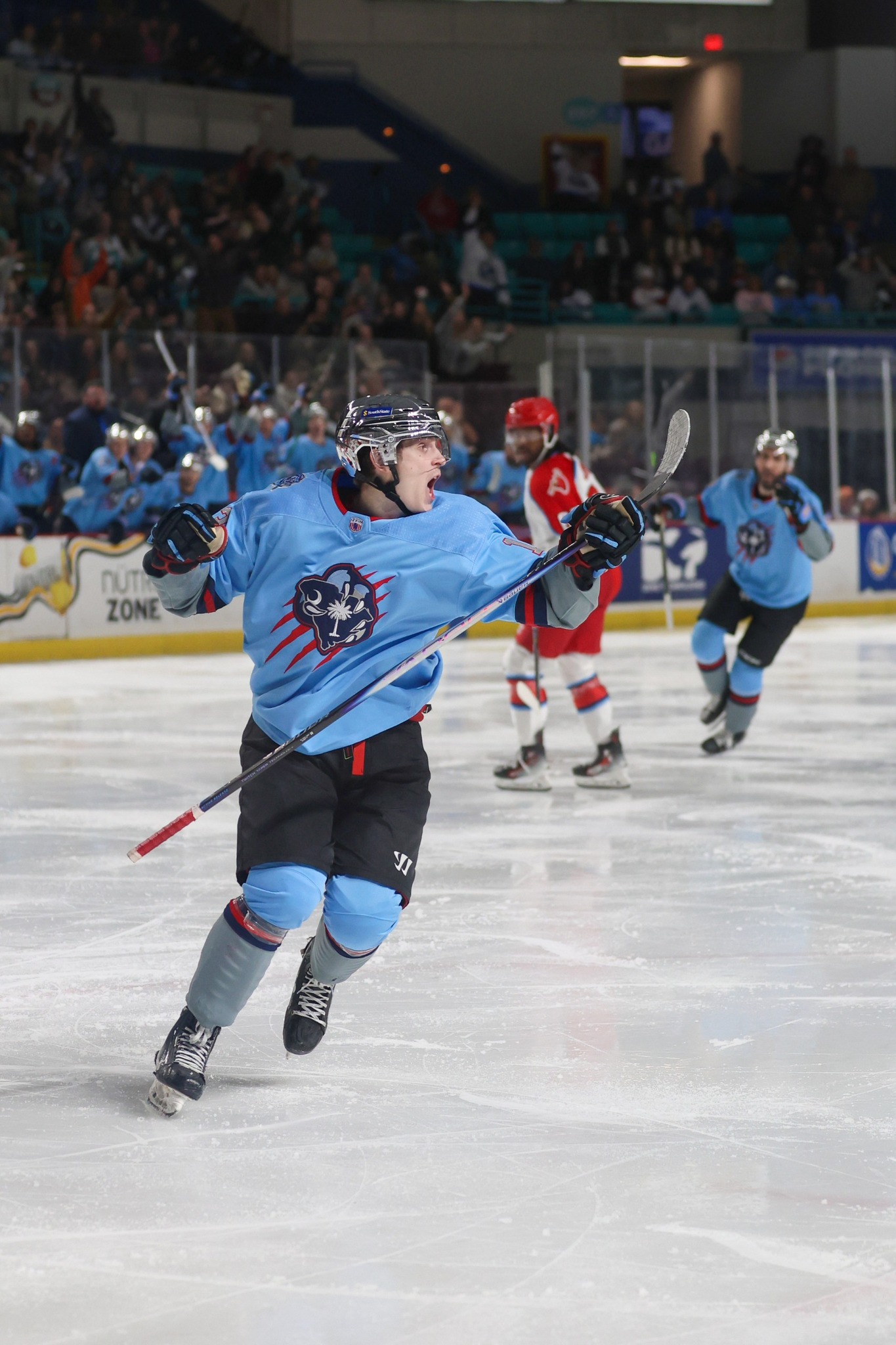 Ice hockey player in a blue uniform celebrates on the ice during a game, teammates and opponents in the background, with fans watching from the stands.