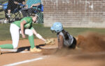 A softball player in green attempts to tag out a sliding runner in grey at home plate, with dust rising from the slide.