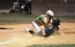 Baseball catcher in green gear tags out a sliding runner in black uniform at home plate, dust flying around them during the play.