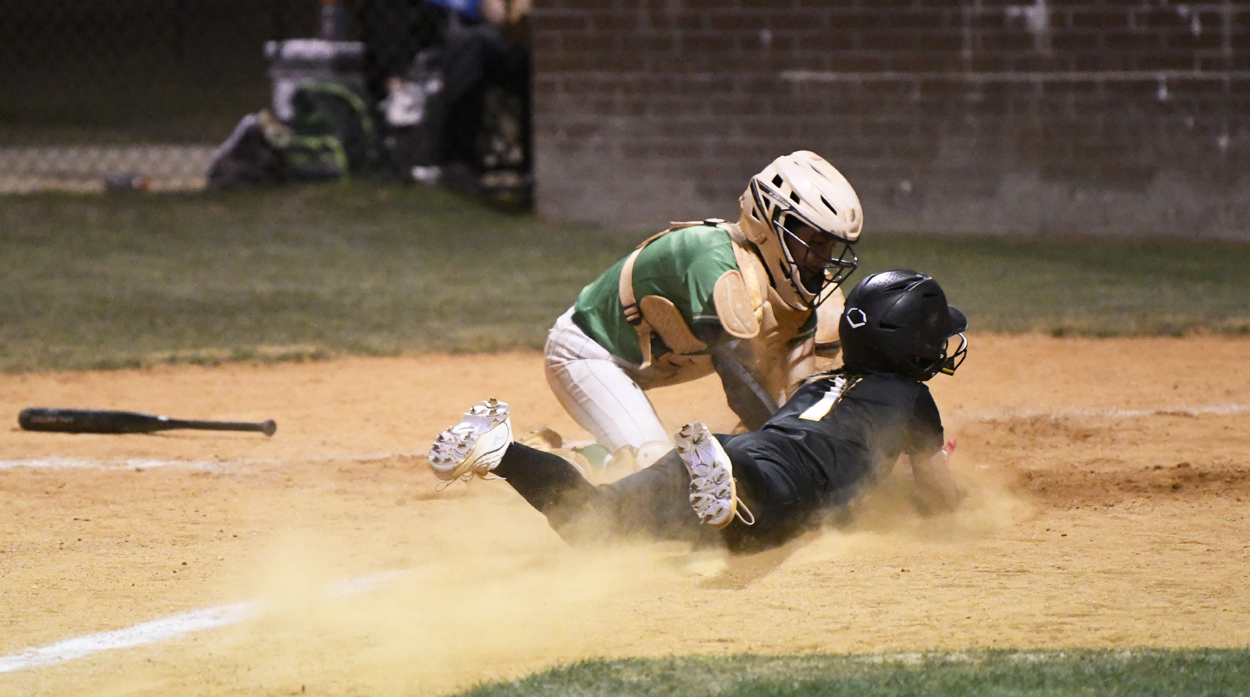 Baseball catcher in green gear tags out a sliding runner in black uniform at home plate, dust flying around them during the play.