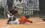 A baseball player in an orange jersey slides into home plate as a catcher attempts to tag him out, with spectators in the background.