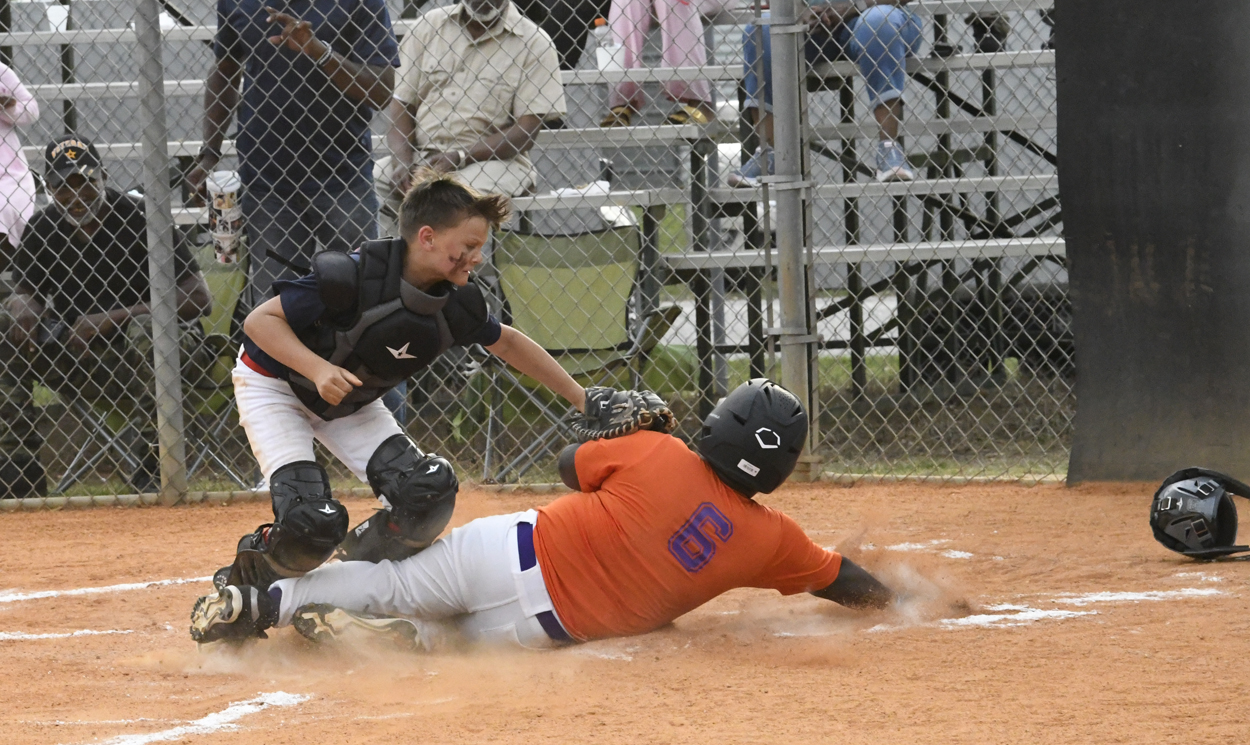 A baseball player in an orange jersey slides into home plate as a catcher attempts to tag him out, with spectators in the background.