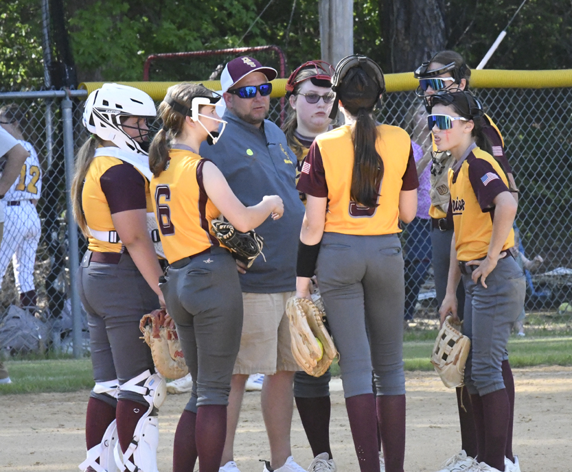 A softball coach talks to players in yellow and maroon uniforms on the field during a game.