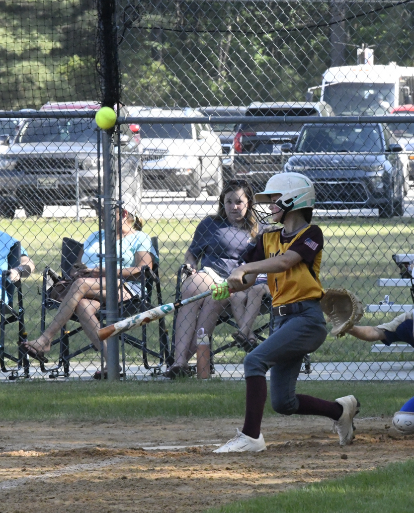 A girl in a yellow jersey and helmet hits a softball with a bat during a game, with spectators and parked cars in the background.