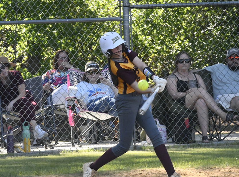A softball player in a yellow and black uniform hits a ball, with spectators watching behind a chain-link fence.