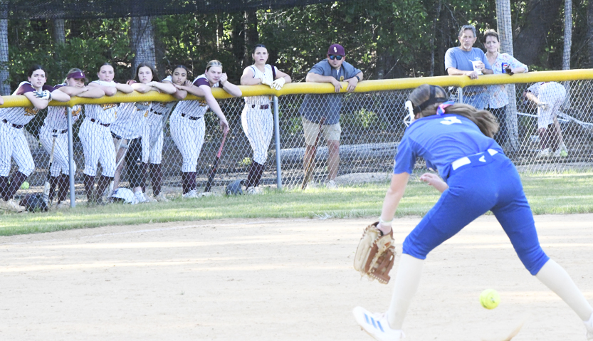 Softball player in blue prepares to field a ball while teammates and coach watch from behind the yellow outfield fence.