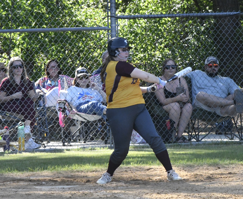 A softball player in yellow and black swings at a pitch while spectators watch from behind a chain-link fence on a sunny day.
